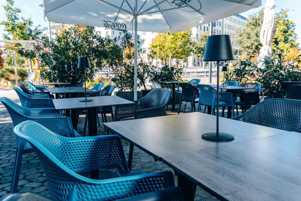 Außencafé Schanigarten mit leeren blauen Stühlen und Holztischen, beschattet von großen Sonnenschirmen und umgeben von Grün an einem sonnigen Tag. Schwarze Tischlampen stehen auf den Tischen.