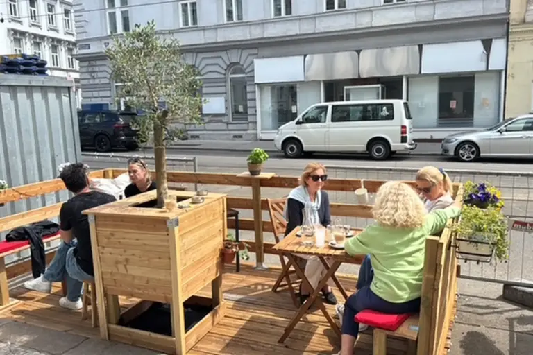Vier Personen sitzen an zwei Holztischen auf einer kleinen Terrasse, die von hölzernen Pflanzkübeln und Bänken umgeben ist. Die Terrasse liegt direkt an einer Straße mit geparkten Autos und Gebäuden im Hintergrund.