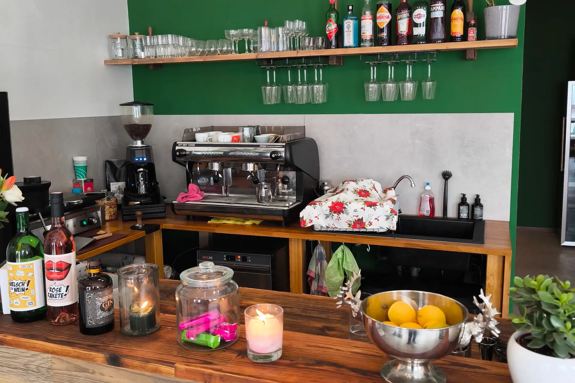 Eine gemütliche Café-Theke mit einer Kaffeemaschine, Glaswaren, Flaschen in Regalen, Kerzen, einer Schale mit Zitronen und bunten Gegenständen in Gläsern, vor einer grünen Wand mit Holzakzenten.
