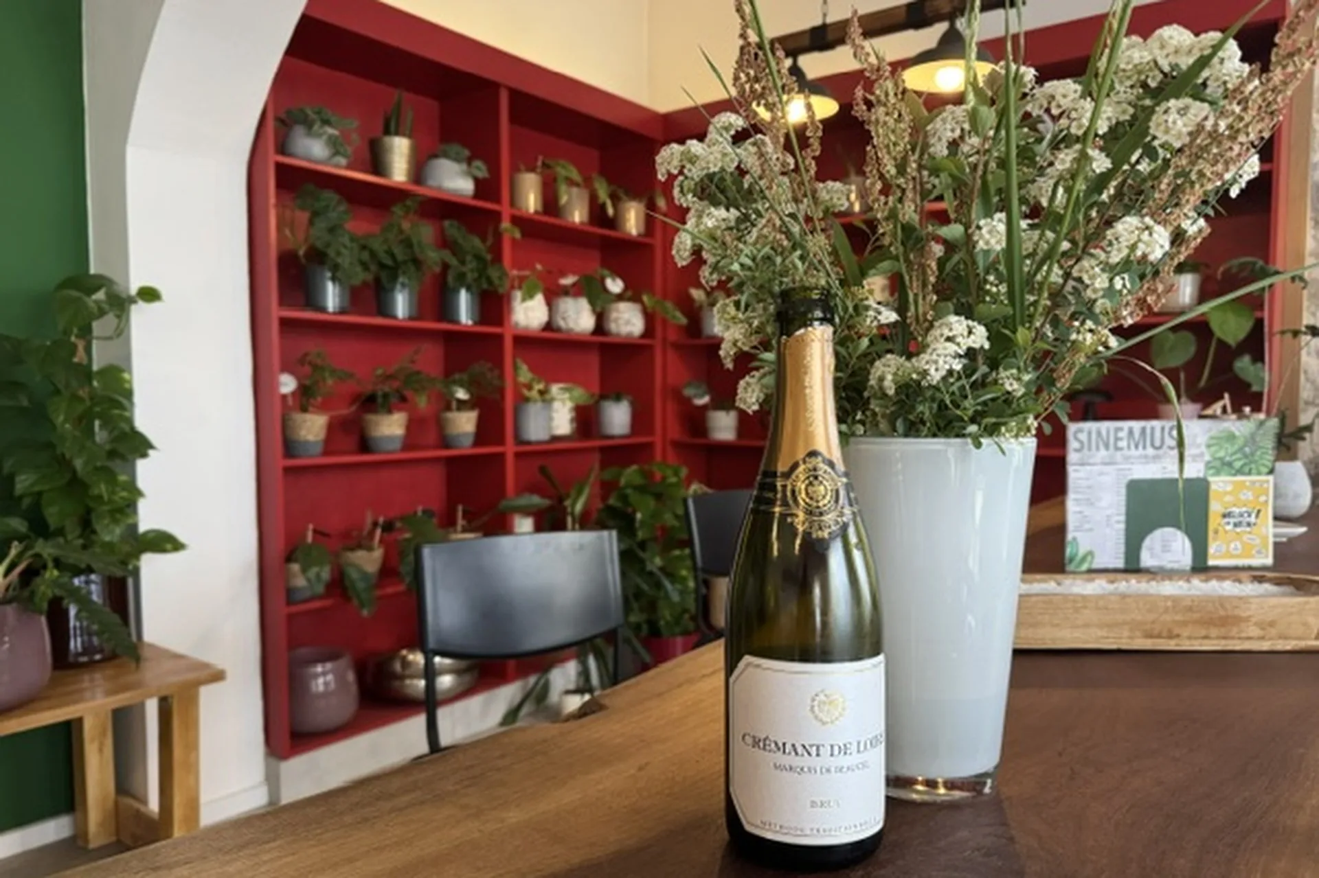 Eine Flasche Sekt mit der Aufschrift "Crémant de Loire" steht auf einem Holztisch neben einer hohen Vase mit weißen Blumen, im Hintergrund stehen Regale mit Topfpflanzen und roten Akzenten.