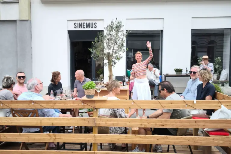 Eine Gruppe von Menschen sitzt an Holztischen vor einem Café namens SINEMUS, redet und trinkt. Eine lächelnde Frau steht und winkt in die Kamera. Pflanzen schmücken die Umgebung, und es scheint ein heller, angenehmer Tag zu sein.