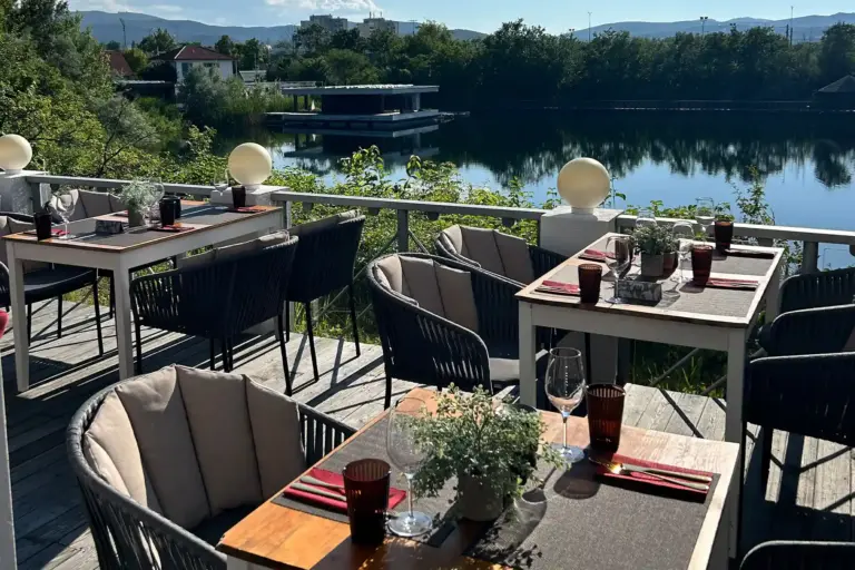 Restaurantterrasse im Freien mit gedeckten Tischen, gepolsterten Stühlen und Gläsern, mit Blick auf einen ruhigen Fluss, umgeben von üppigem Grün und entfernten Gebäuden bei klarem, sonnigem Himmel.