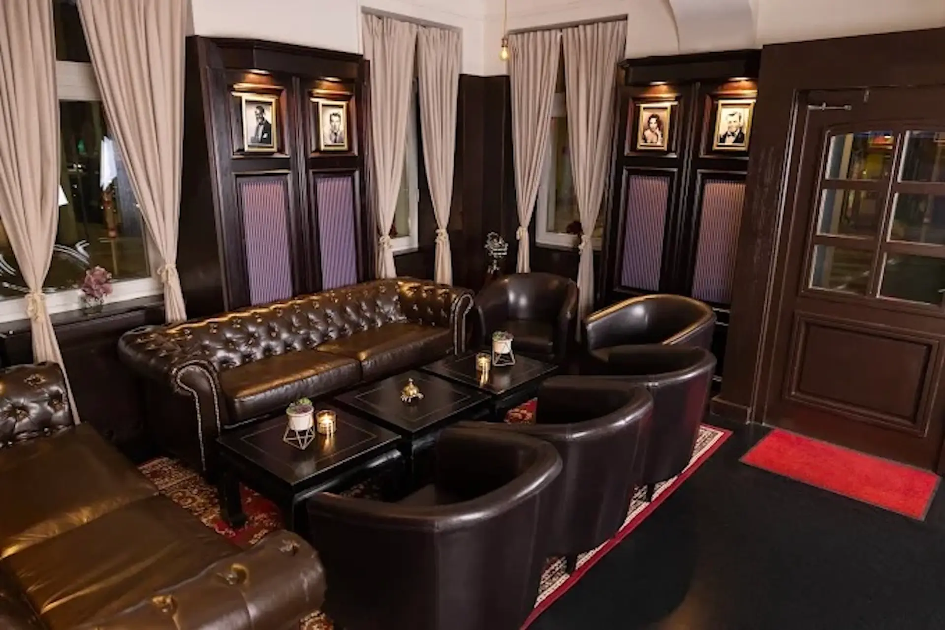 Eine gemütliche Lounge mit dunklen Ledersesseln und einem getufteten Sofa, die um niedrige schwarze Tische angeordnet sind; gerahmte Fotos an getäfelten Wänden, hohe Vorhänge, sanfte Beleuchtung und ein roter Teppich schaffen eine warme, einladende Atmosphäre.
