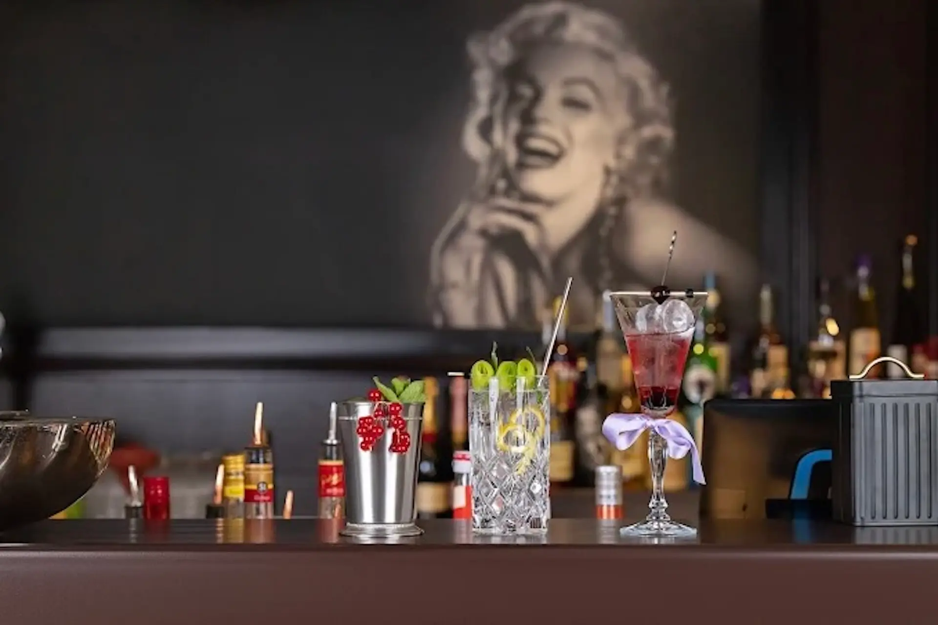 Ein Bartresen mit drei bunten Cocktails, Flaschen und Mixern im Hintergrund, dahinter das große Schwarz-Weiß-Porträt einer lächelnden Frau an der Wand.