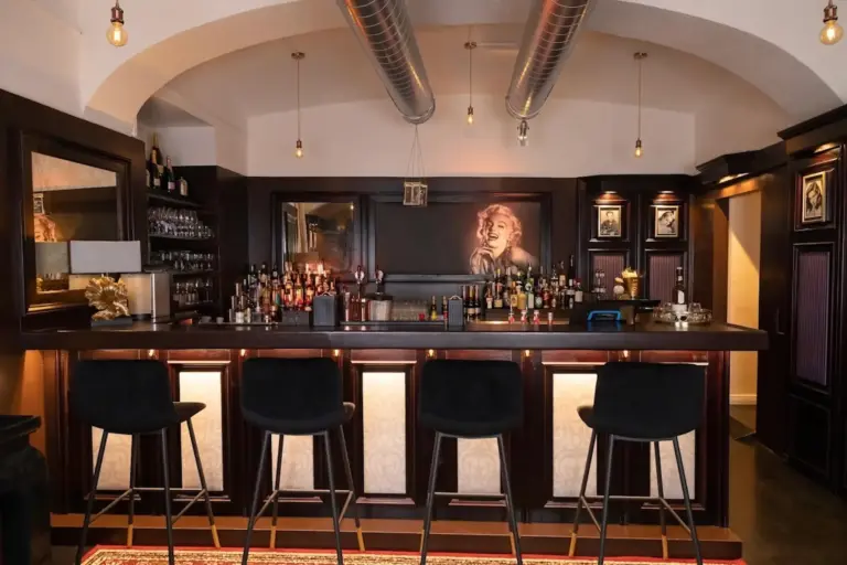 Eine stilvolle Bar mit vier schwarzen Hockern, einem dunklen Holztresen, Flaschenregalen und einem großen Porträt einer Frau mit hellem Haar hinter der Bar. Von der Decke hängen Hängelampen und metallene Luftkanäle.