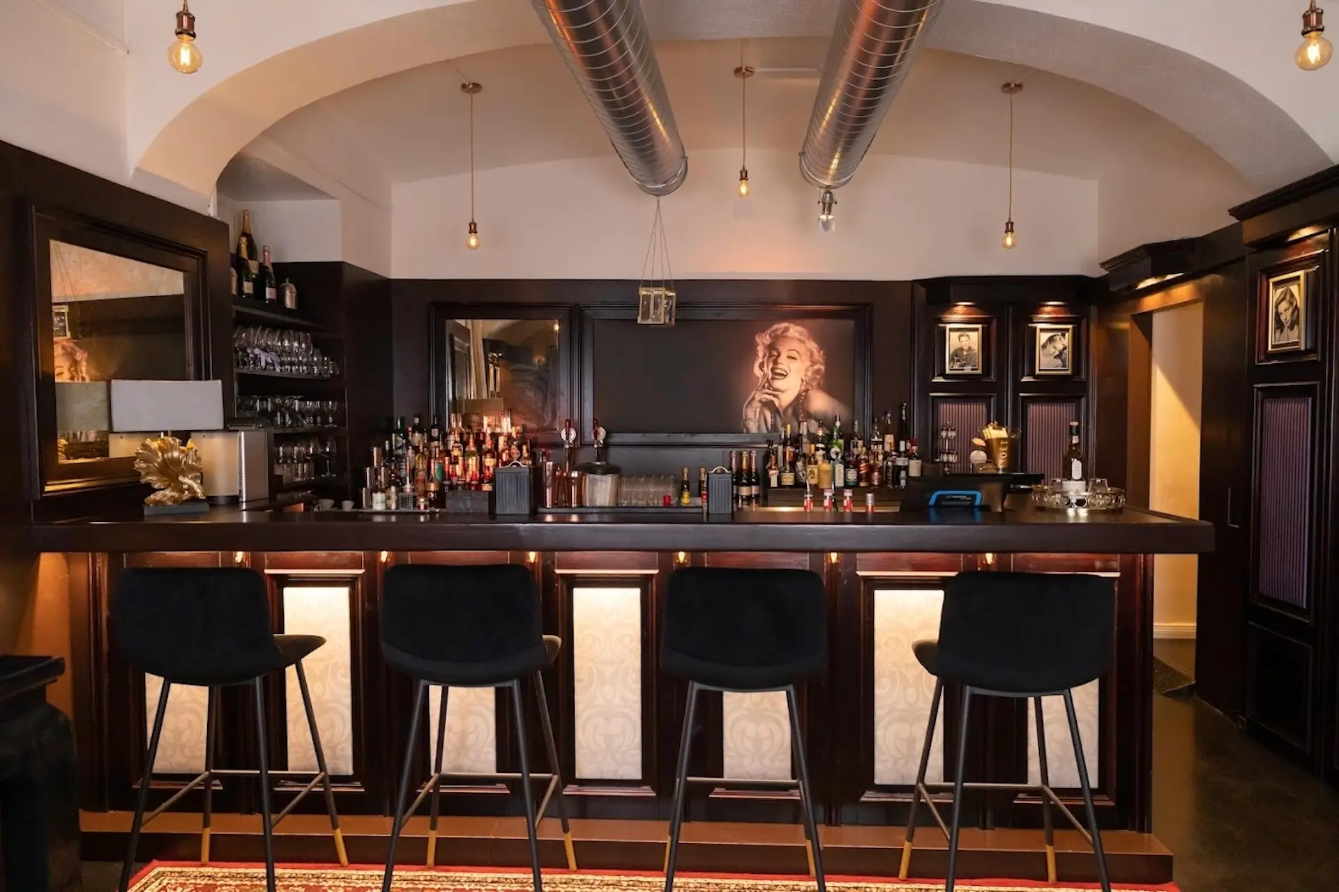 Eine stilvolle Bar mit vier schwarzen Hockern, einem dunklen Holztresen, Flaschenregalen und einem großen Porträt einer Frau mit hellem Haar hinter der Bar. Von der Decke hängen Hängelampen und metallene Luftkanäle.