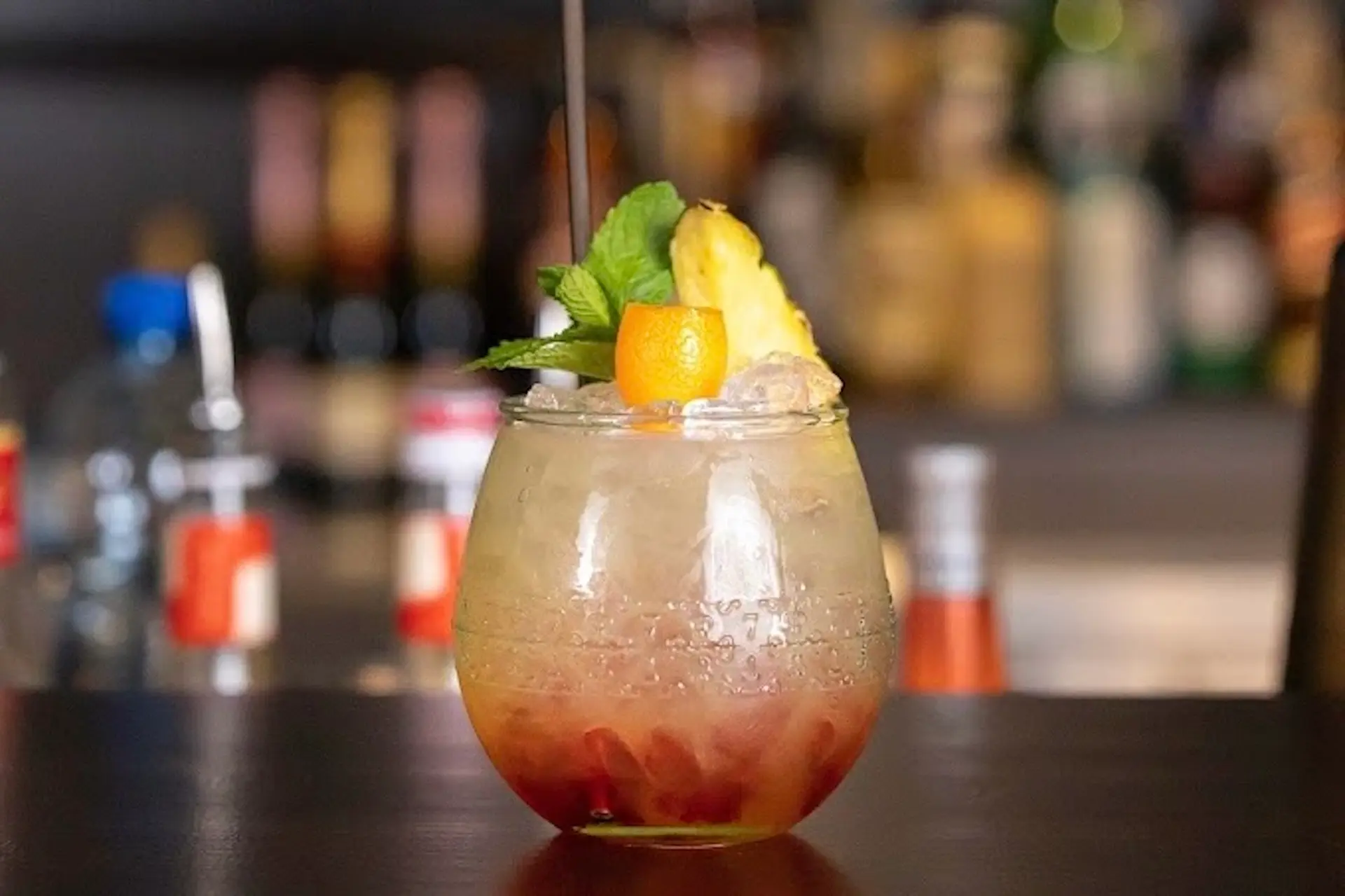 Ein rundes Glas, gefüllt mit einem bunten Cocktail, garniert mit einem Minzzweig, einer Orangenschale, einer Ananasscheibe und einem schwarzen Strohhalm, steht auf einer dunklen Bar mit verschwommenen Flaschen im Hintergrund.