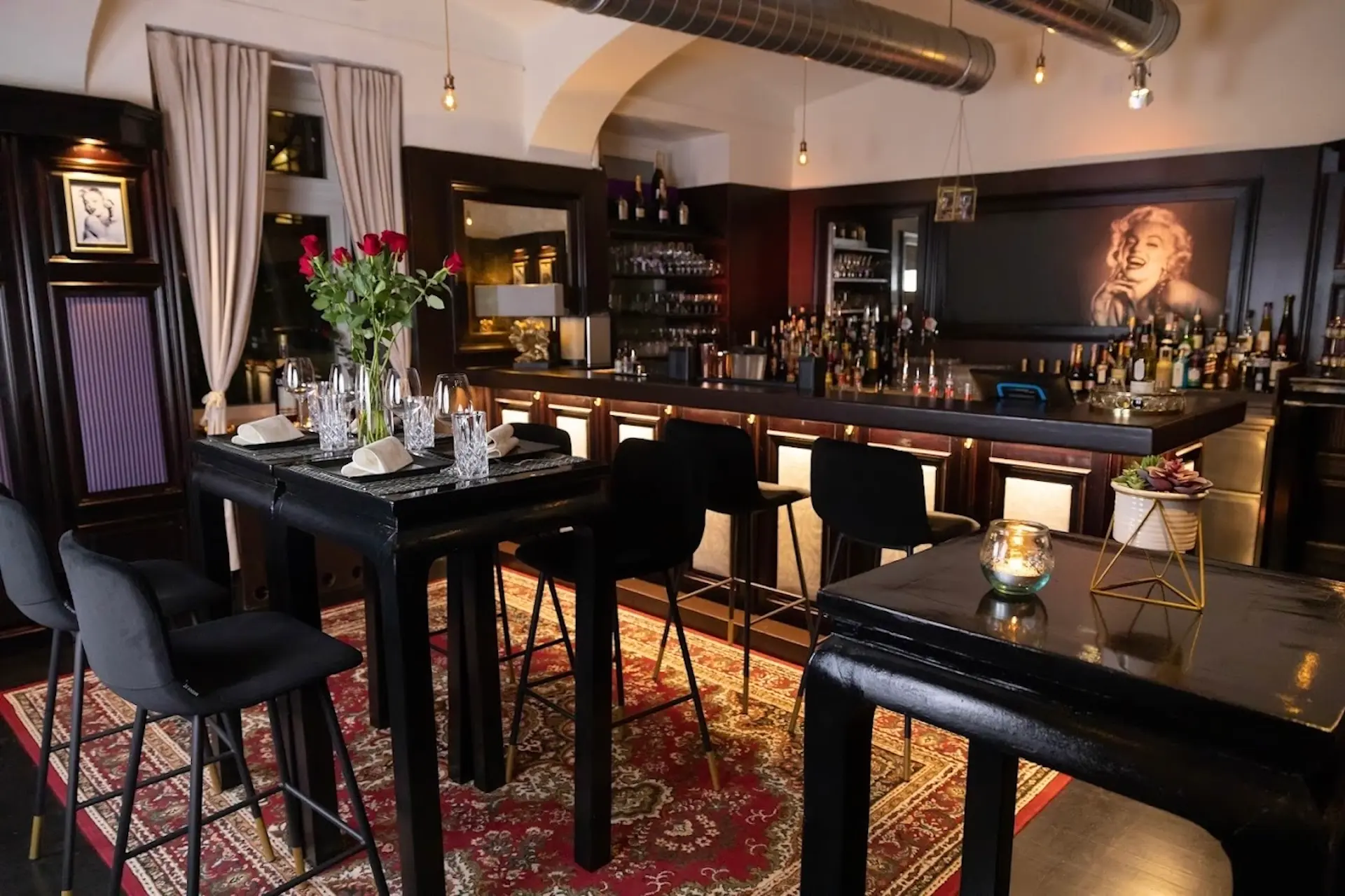 Schickes Bar-Interieur mit hohen Tischen, schwarzen Stühlen, roten Rosen in einer Vase, gedämpfter Beleuchtung, einem gemusterten Teppich und einem Porträt von Marilyn Monroe an der Wand hinter einer gut bestückten Bar.