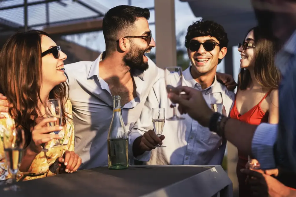 Vier junge Erwachsene mit Sonnenbrillen lachen und unterhalten sich auf einem Sommerfest in Wien, halten Sektgläser in der Hand und haben eine Flasche Sekt vor sich auf dem Tisch stehen.