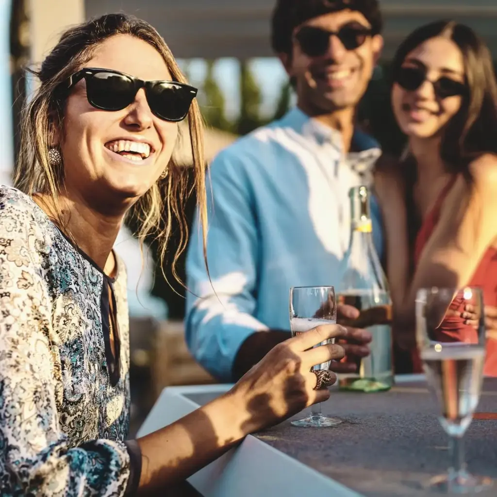 Drei Personen mit Sonnenbrillen lächeln und trinken gemeinsam an einem sonnenbeschienenen Tisch während eines Sommerfestes in Wien, wobei eine Frau im Vordergrund ein Glas hält und lacht.