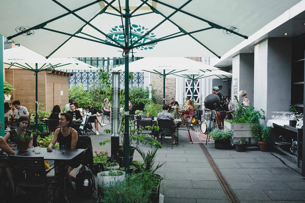 Man sitzt und speist an Tischen im Freien unter großen weißen Sonnenschirmen auf einer belebten Schanigarten-Terrasse, umgeben von Topfpflanzen und Grünzeug, mit einem modernen Gebäude im Hintergrund.