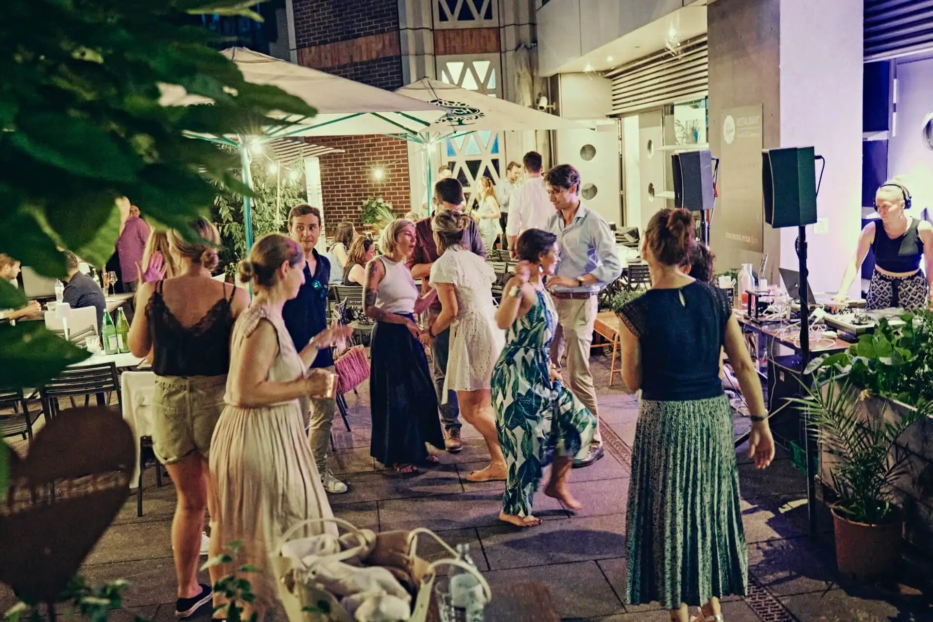 Eine Gruppe sommerlich gekleideter Menschen tanzt und unterhält sich auf einer mit Pflanzen und Lichterketten geschmückten Abendparty im Freien. Einige unterhalten sich, während andere tanzen, was eine lebhafte und festliche Atmosphäre schafft.