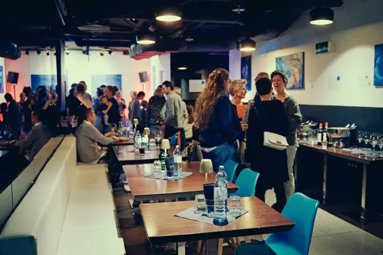 Eine lebhafte Restaurantszene mit Gruppen von Menschen, die sich unterhalten und an Tischen stehen. Die Tische sind mit Wasserflaschen, Gläsern und Servietten gedeckt. Die Beleuchtung ist gedämpft und die Atmosphäre wirkt geschäftig und lebendig.