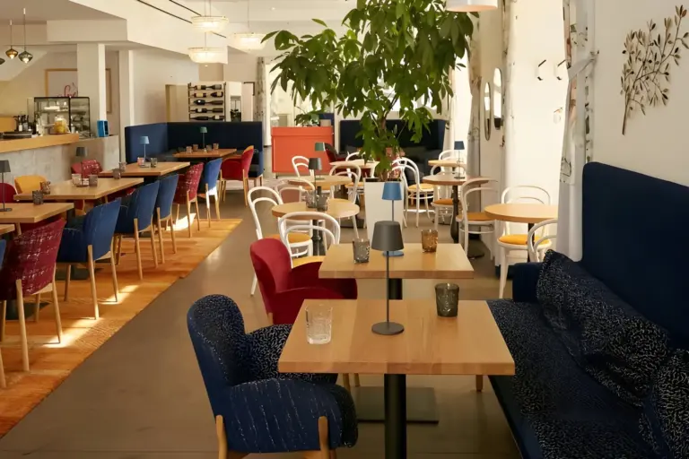 Ein helles, modernes Restaurant mit Holztischen, bunten Stühlen, blauen Sofas und Topfbäumen. Der Raum ist durch große Fenster und stilvolle Pendelleuchten gut beleuchtet und schafft eine einladende Atmosphäre.