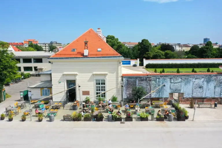 Luftaufnahme eines Straßencafés mit gelben Stühlen und Tischen, Menschen sitzen und entspannen sich, viele Topfpflanzen, neben einem weißen Gebäude mit rotem Dach, unter einem klaren blauen Himmel in einer städtischen Umgebung.