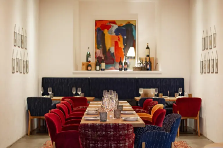 Ein gemütlicher Essbereich in einem Restaurant mit einem langen Holztisch, bunten Samtstühlen, Weingläsern auf dem Tisch und moderner Wandkunst über einem Regal mit Weinflaschen. Die warme Beleuchtung schafft eine einladende Atmosphäre.
