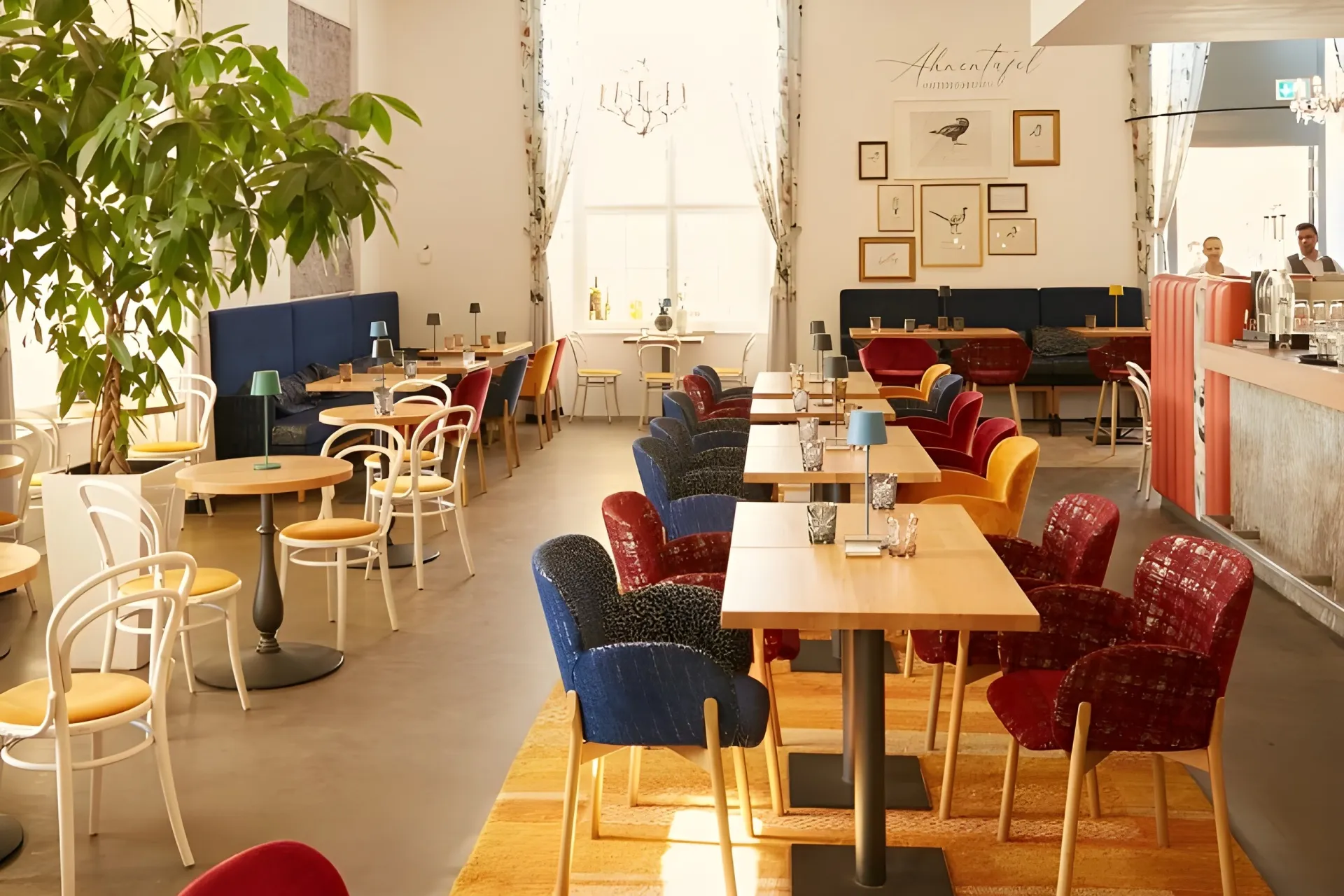 Ein helles, modernes Café mit Holztischen, bunten Samtstühlen und weißen Stühlen. Sonnenlicht strömt durch ein großes Fenster, und Kunstwerke schmücken die Wände. Grünpflanzen und Hängelampen tragen zur gemütlichen Atmosphäre bei.