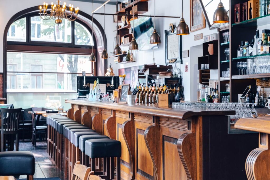 Eine stilvolle, leere Bar mit Holzhockern, einem langen, polierten Tresen, Regalen mit Flaschen und Gläsern, Pendelleuchten und großen Bogenfenstern, die natürliches Licht hereinlassen.
