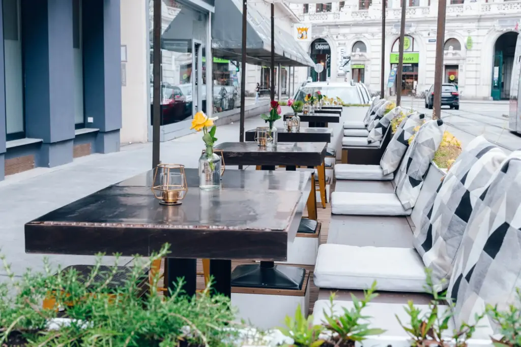 Café im Freien mit dunklen Holztischen, gepolsterten Stühlen und kleinen Blumenvasen auf jedem Tisch. Der Bereich ist mit Pflanzen gesäumt und bietet einen Blick auf eine ruhige Stadtstraße mit Gebäuden im Hintergrund.