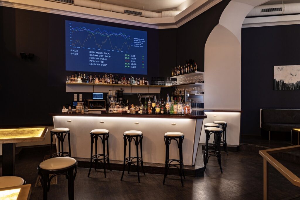 Stage Bar Eine moderne Stage Bar mit einem geschwungenen Tresen, mehreren Barhockern und verschiedenen Flaschen in der Auslage. Ein großer Bildschirm über der Bar zeigt bunte Finanzdiagramme vor einer dunklen Wand und schafft eine warme und doch anspruchsvolle Atmosphäre in dem leeren Raum.