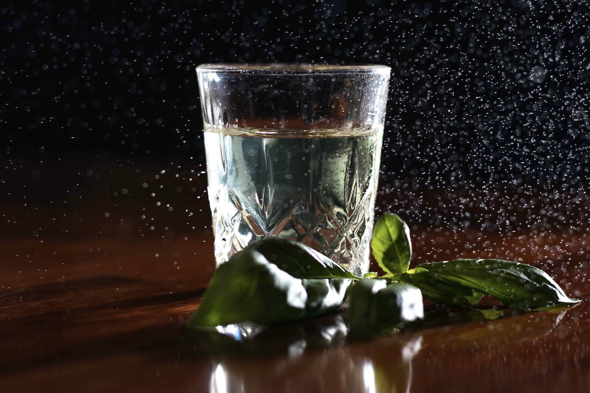 Auf einer dunklen, spiegelnden Fläche an der Stage Bar steht ein Glas Wasser, dessen Tropfen in der Luft schweben. Grüne Basilikumblätter liegen vor dem Glas, während ein schwarzer Hintergrund den dramatischen Effekt noch verstärkt.