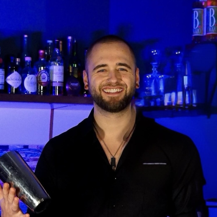 Ein lächelnder Barkeeper mit Bart und rasiertem Kopf steht hinter der Stage Bar und hält einen Cocktailshaker in einer Hand. Im Hintergrund sind unter blauer Beleuchtung Regale mit verschiedenen Flaschen zu sehen.