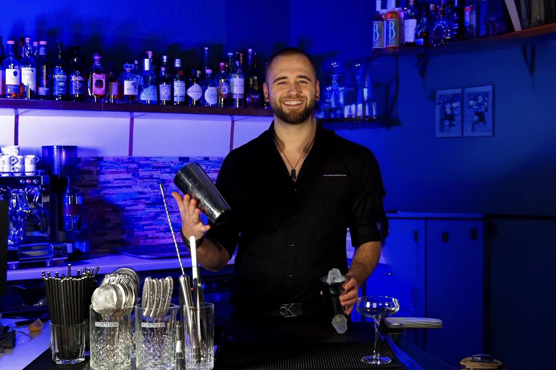 Ein lächelnder Barkeeper in einem schwarzen Hemd hält einen Cocktailshaker und einen Jigger hinter der Stage Bar, umgeben von Barwerkzeugen, Gläsern und Regalen mit Flaschen, alles unter blauer Beleuchtung.