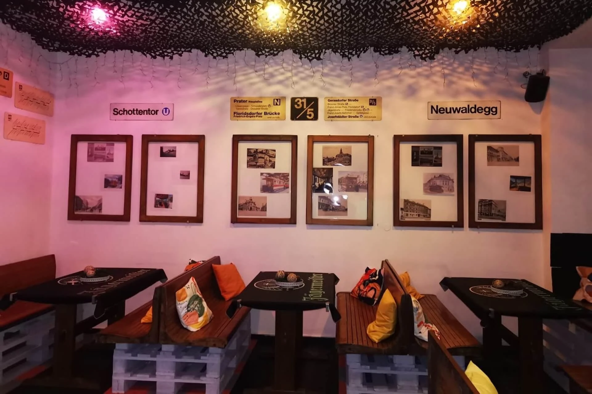 Gemütliche Sitzecke im Innenbereich mit Holzbänken, Paletten als Unterlage, bunten Kissen und gerahmten Fotos an der Wand. Darüber hängen Schilder im U-Bahn-Stil. Warme Beleuchtung und eine Netzdecke schaffen eine einladende Atmosphäre.