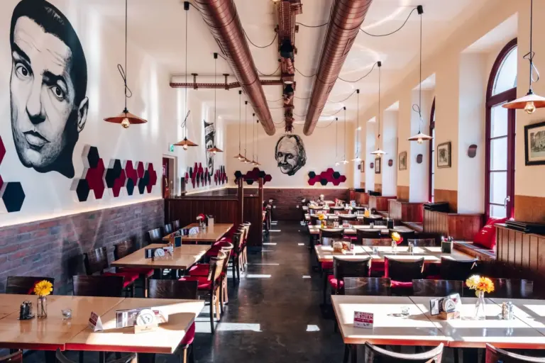 Ein geräumiges Restaurant mit Holztischen und roten Stühlen, dekoriert mit großen schwarz-weißen Wandbildern von Gesichtern an den Wänden, Hängelampen und sechseckiger Wandkunst. Das Sonnenlicht strömt durch die hohen Fenster.