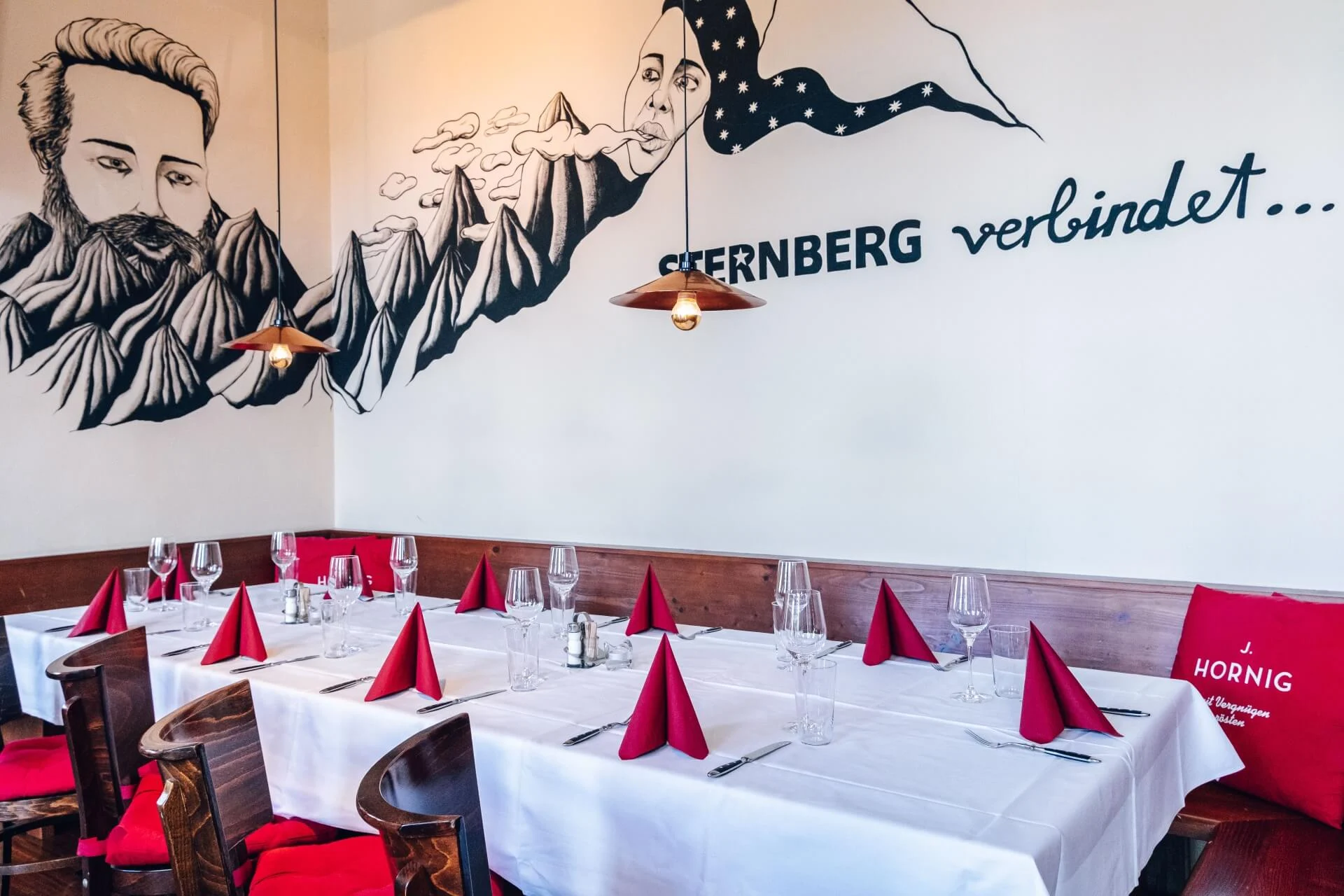 Ein mit weißen Tüchern, Gläsern und roten Servietten gedeckter Tisch in einem gemütlichen Essbereich. An der Wand befinden sich Wandmalereien mit Bergmotiven und der Aufschrift "Sternberg verbindet..." über den Sitzplätzen.