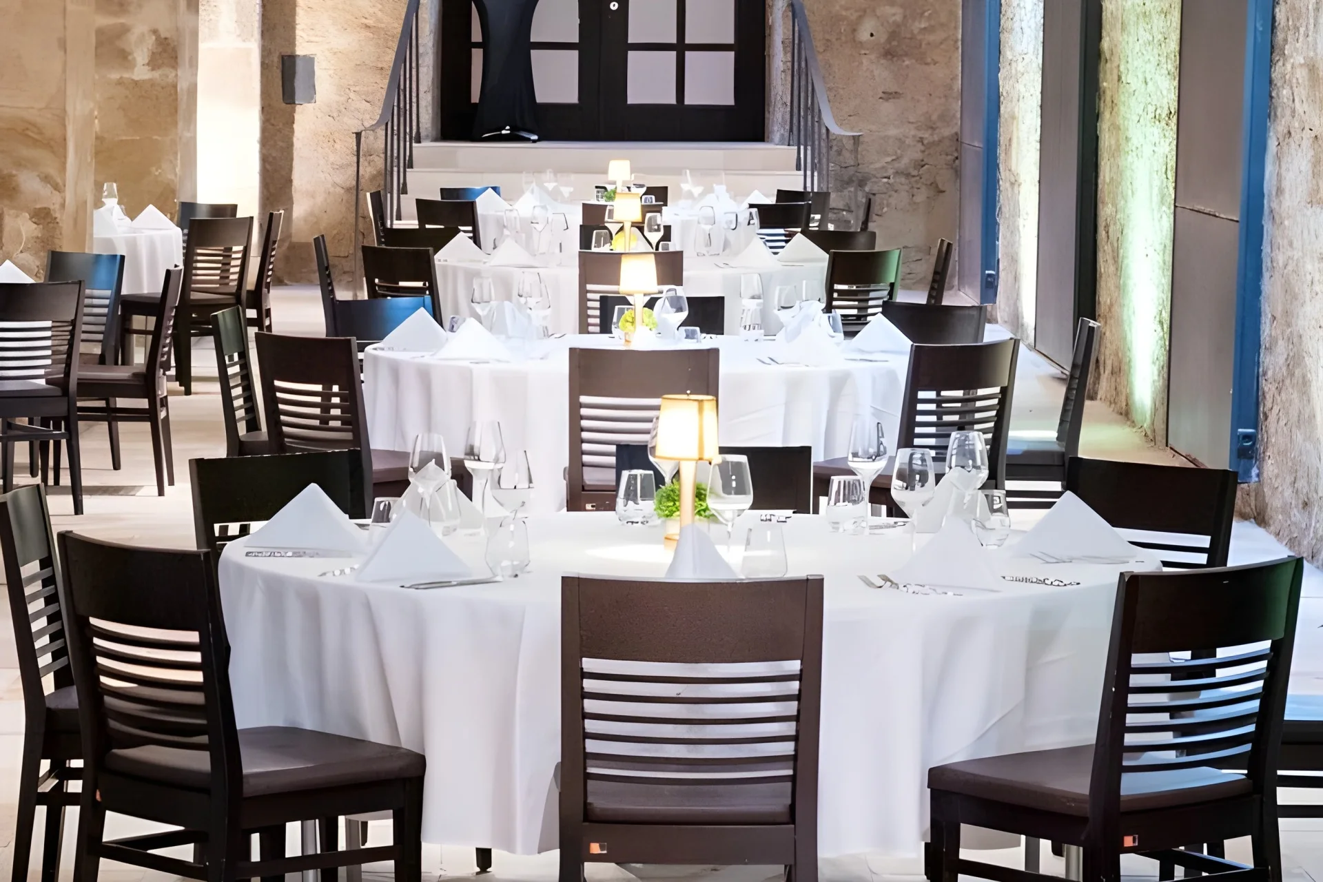 Elegantes Restaurantinterieur mit runden Tischen mit weißen Tischtüchern, ordentlich angeordneten Gedecken, Weingläsern, Servietten und einer kleinen Lampe in der Mitte, umgeben von dunklen Holzstühlen. Steinmauern und Treppen im Hintergrund.