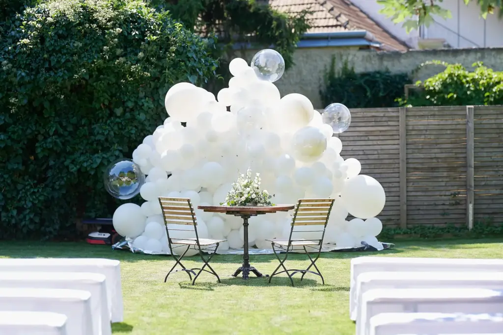 Zwei Holzstühle und ein runder Tisch mit einem Blumenschmuck stehen auf dem Rasen vor einer großen Anzahl weißer Luftballons im Freien, mit Bänken im Vordergrund und Grünpflanzen im Hintergrund.