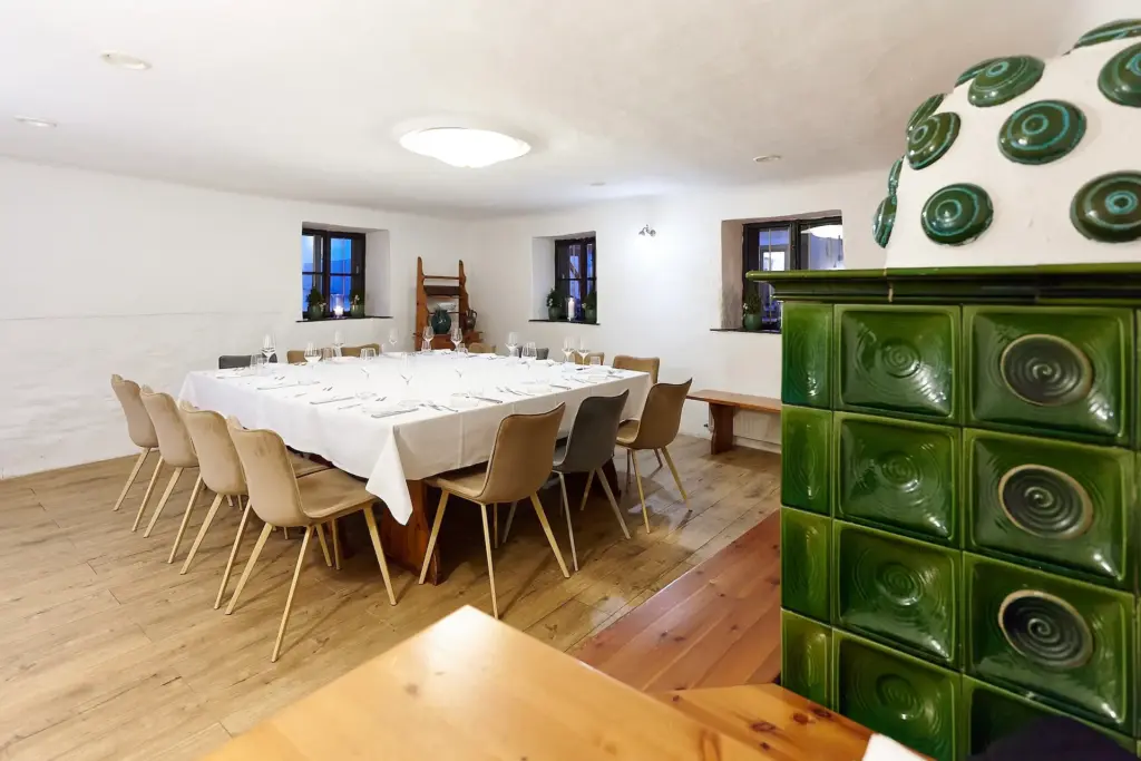 Ein Esszimmer mit einem langen Tisch, der für eine Mahlzeit gedeckt ist, umgeben von Holzstühlen. Der Raum hat weiße Wände, Holzböden, kleine Fenster und einen grünen Kachelofen im Vordergrund.