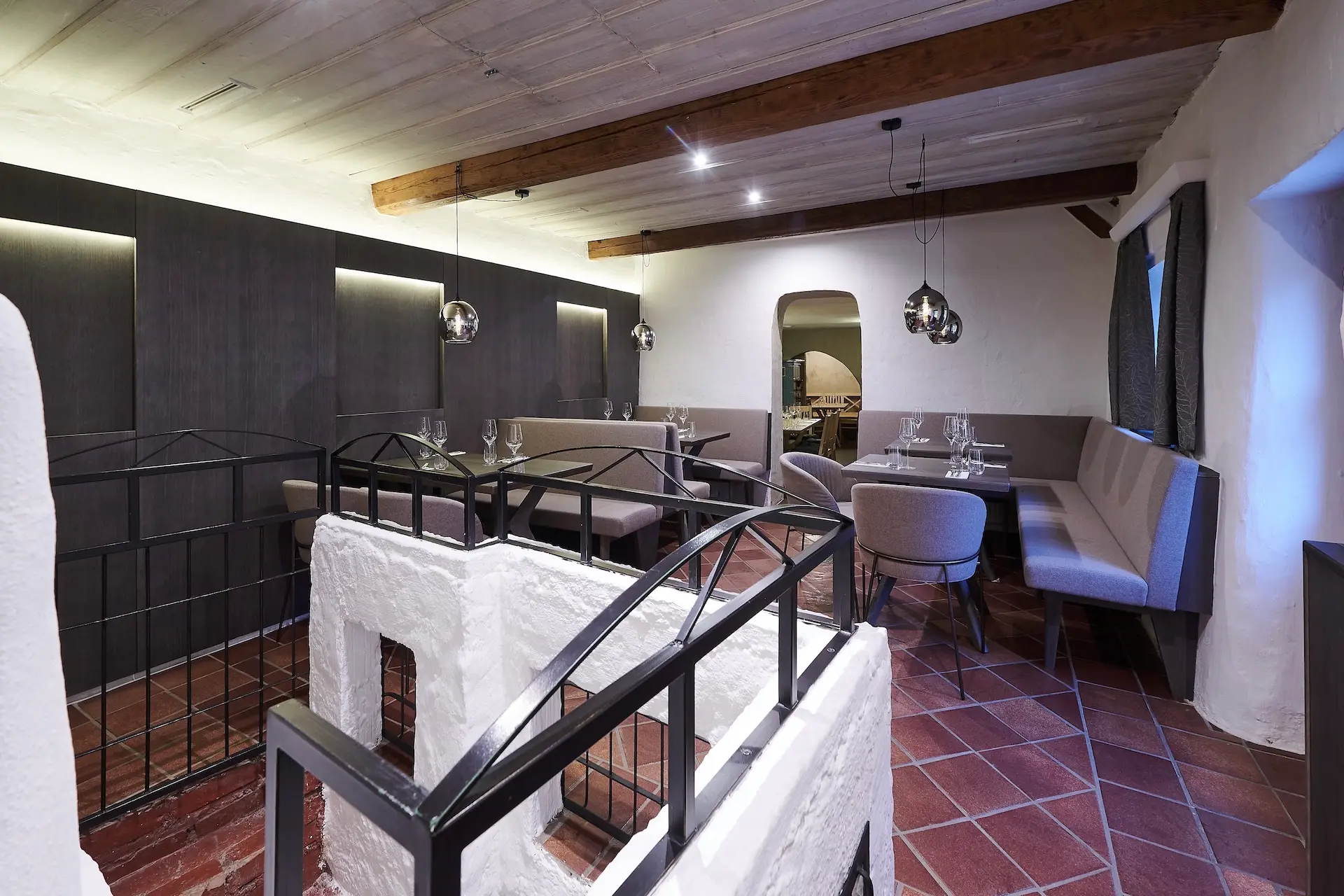 Modernes Restaurantinterieur mit Holzbalken, dunklen Wänden, Hängelampen und Tischen mit Glaswaren. Weißer Putz und schwarze Geländer umgeben eine Treppe im Vordergrund. Der Boden ist mit braunen Fliesen ausgelegt.