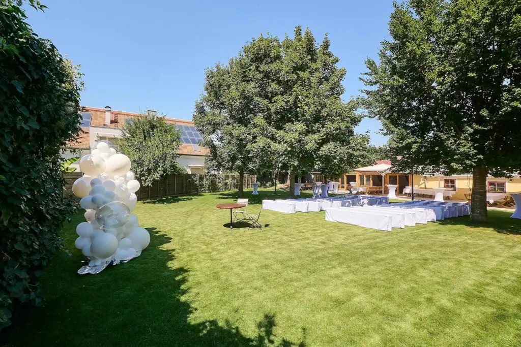 Eine sonnige Veranstaltung im Freien auf einer Wiese mit weißen Luftballons, Reihen von weiß bezogenen Stühlen, Tischen und großen schattenspendenden Bäumen. Im Hintergrund ist ein Gebäude mit Sonnenkollektoren zu sehen.