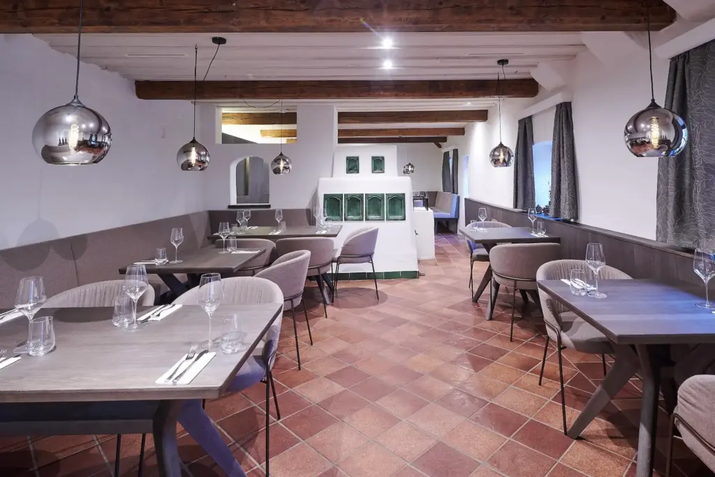 Ein modernes Restaurant mit Holzbalken, Pendelleuchten, gefliestem Boden, grauen Stühlen und gedeckten Tischen mit Gläsern und Besteck, die eine gemütliche und elegante Essatmosphäre schaffen.