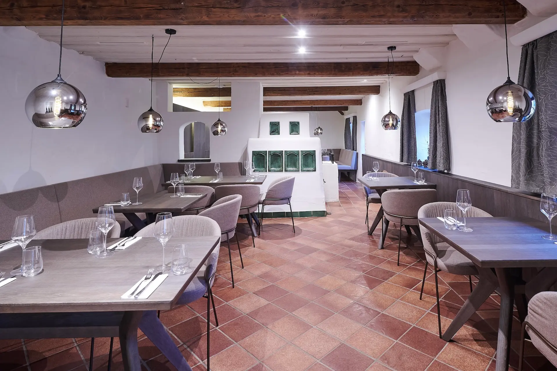 Ein modernes Restaurant mit Holzbalken, Pendelleuchten, gefliestem Boden, grauen Stühlen und gedeckten Tischen mit Gläsern und Besteck, die eine gemütliche und elegante Essatmosphäre schaffen.
