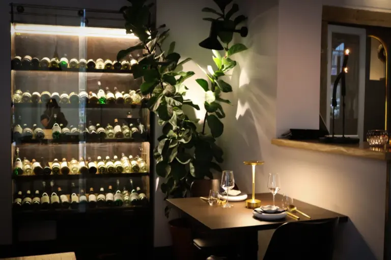 Eine gemütliche Restaurantecke mit einem gedeckten Tisch für zwei Personen, einer goldenen Lampe, Weingläsern, Tellern und Besteck. Hinter dem Tisch befinden sich ein großes Weinregal und eine hohe Blattpflanze, die eine einladende, intime Atmosphäre schaffen.