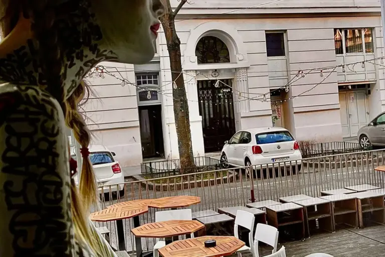 Eine Schaufensterpuppe mit tätowierungsähnlichen Motiven steht an einem Fenster und blickt auf leere Tische und Stühle eines Straßencafés. Auf der anderen Straßenseite parken zwei weiße Autos vor einem hellen Gebäude mit Rundbogenfenstern.