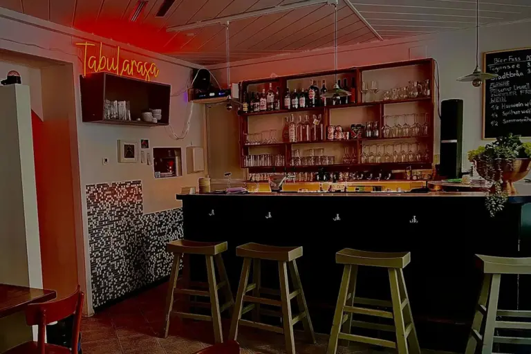 Tabularasa Eine gemütliche Bar mit vier Holzhockern an einem schwarzen Tresen, dahinter Regale mit Flaschen und Gläsern, Mosaikfliesen-Akzente, ein Neonschild von Tabul parasco und eine warme Raumbeleuchtung.