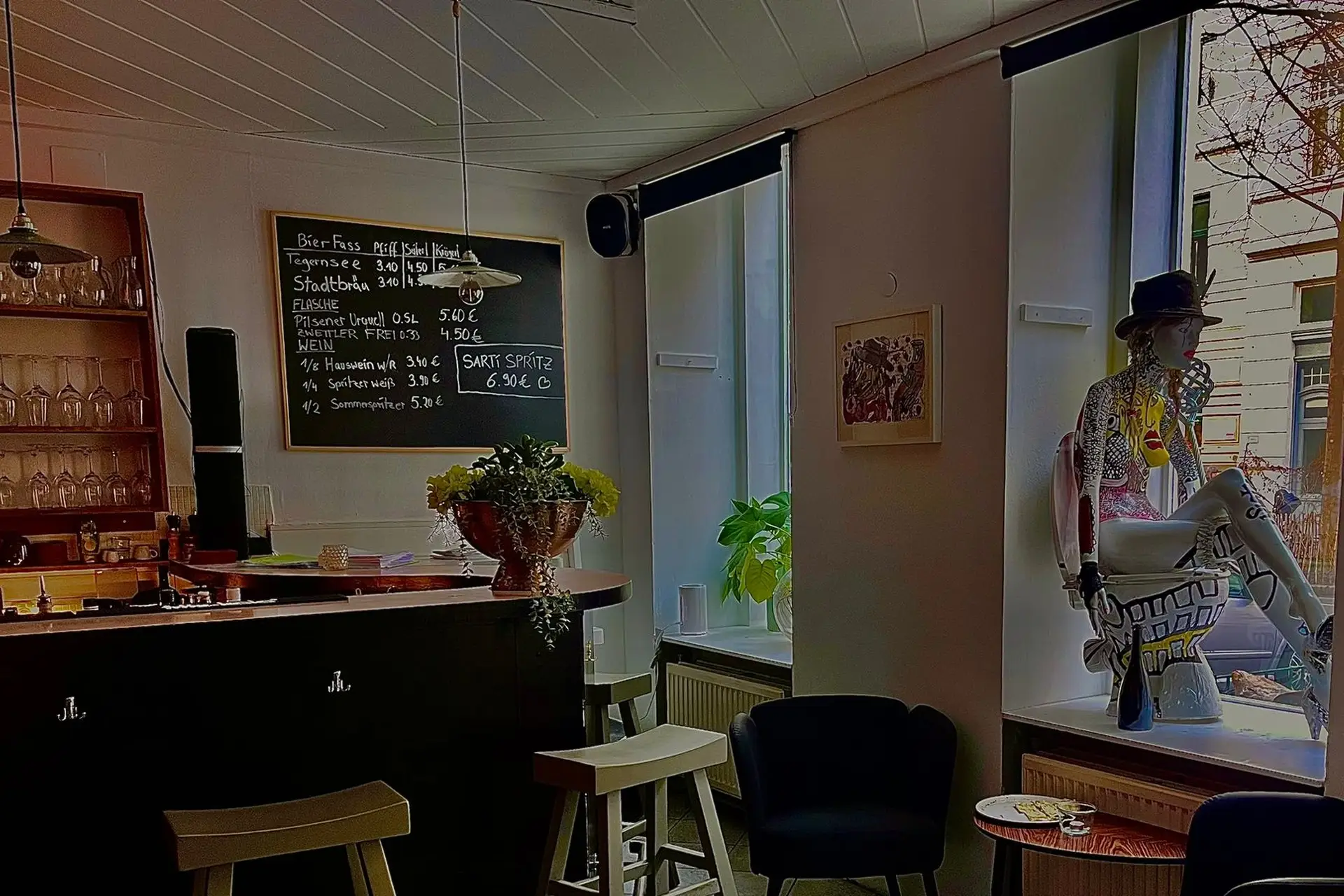 Eine gemütliche Bar mit einem hölzernen Tresen, einer Kreidetafel als Speisekarte, Regalen mit Gläsern, einem kleinen Tisch mit Blumen, modernen Stühlen und einer mit bunten Mustern verzierten Schaufensterpuppe am Fenster.