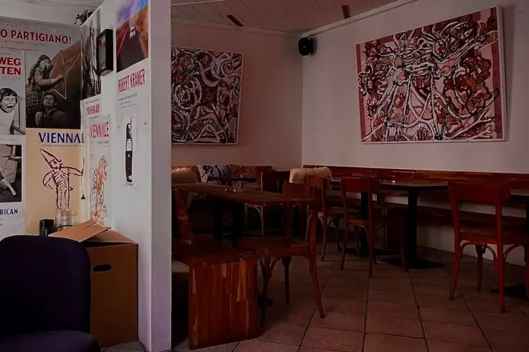 Ein gemütliches Café mit Holztischen und -stühlen, abstrakter Kunst an den Wänden und Postern an einer weißen Säule. Der Boden ist gefliest und der Raum ist sanft beleuchtet, was eine warme und einladende Atmosphäre schafft.