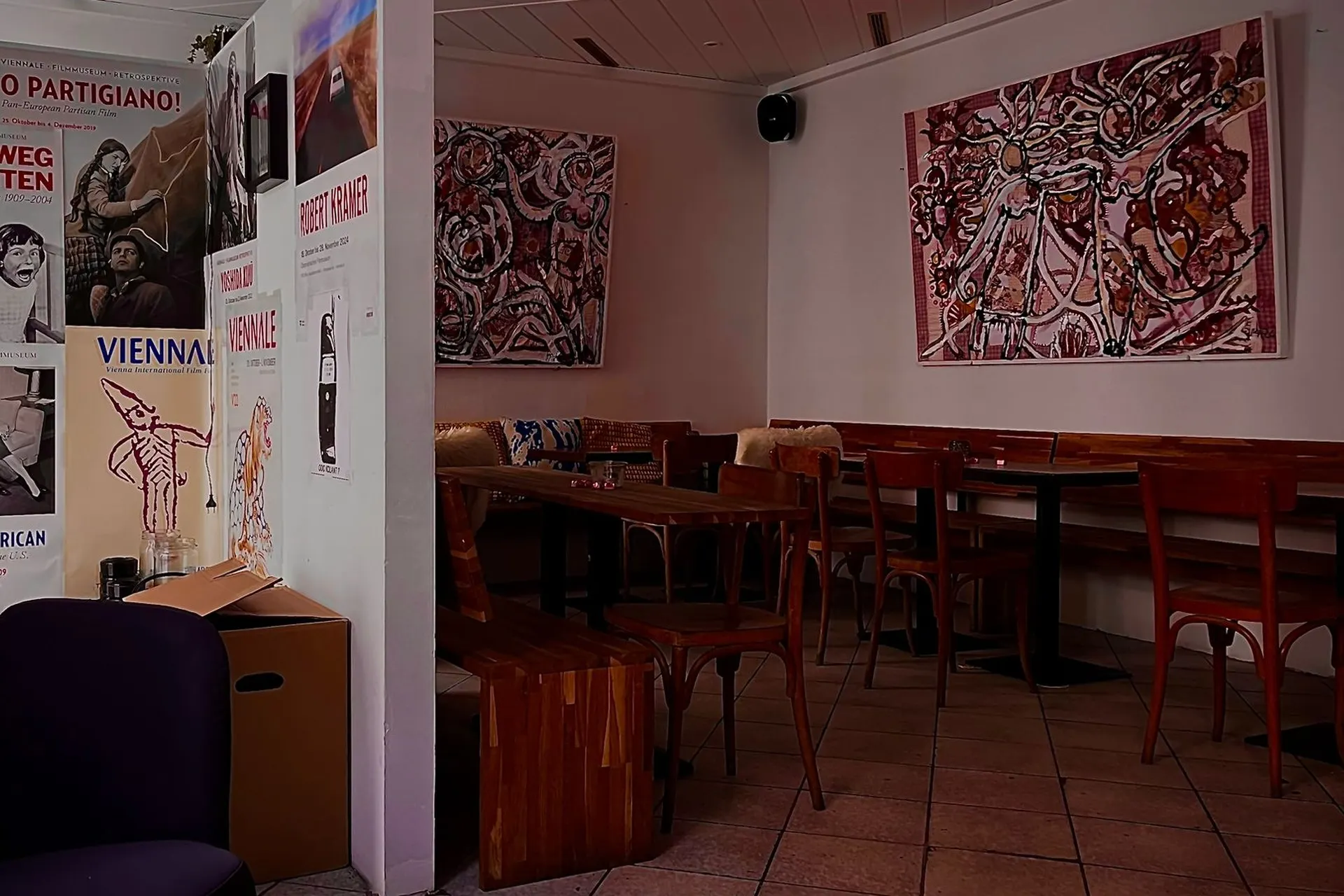 Ein gemütliches Café mit Holztischen und -stühlen, abstrakter Kunst an den Wänden und Postern an einer weißen Säule. Der Boden ist gefliest und der Raum ist sanft beleuchtet, was eine warme und einladende Atmosphäre schafft.