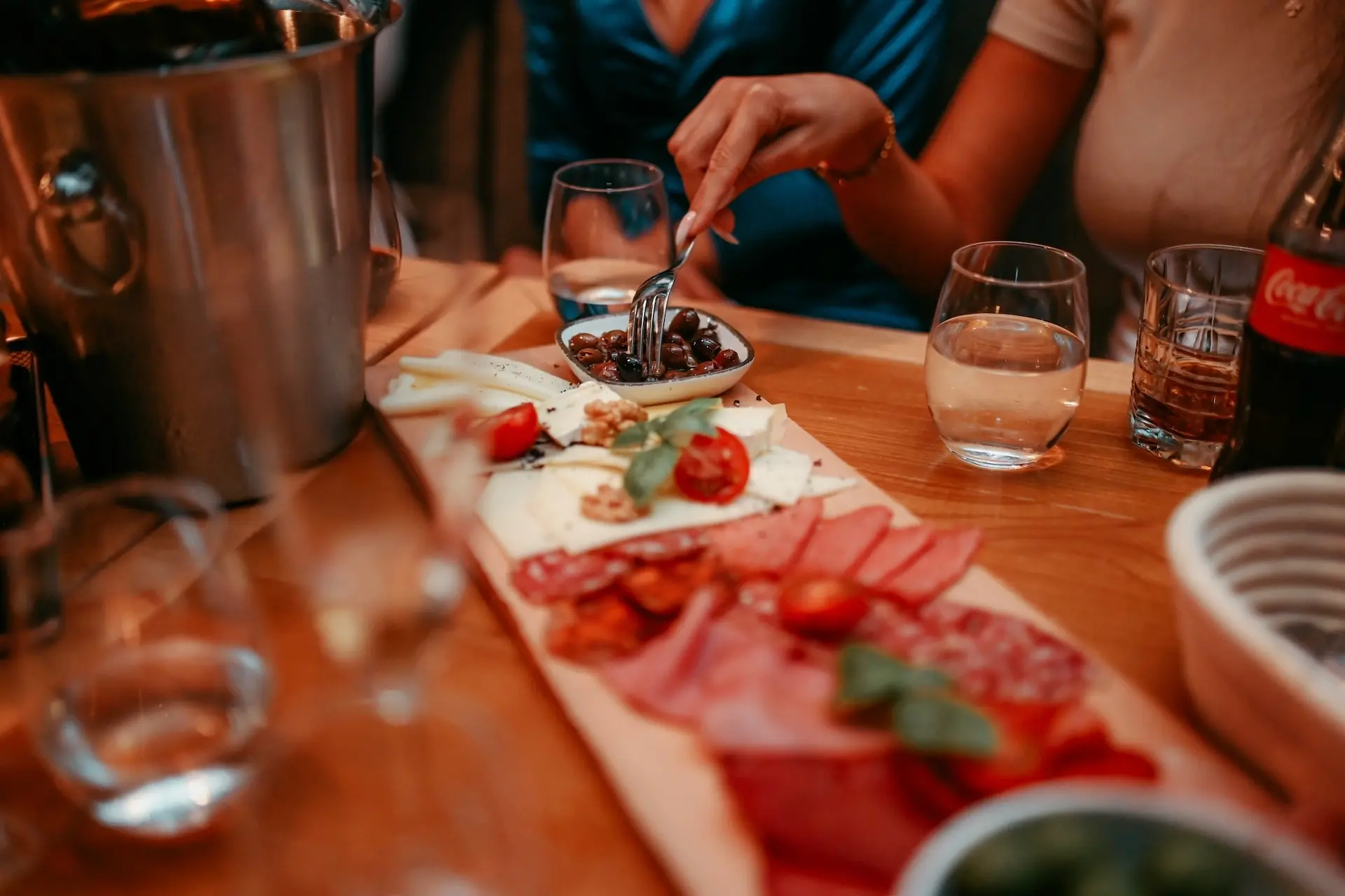 Eine Nahaufnahme eines Wurstbretts mit Fleisch, Käse, Walnüssen und Tomaten auf einem Tisch. Zwei Personen sitzen, eine greift nach einer Schale mit Oliven. Gläser mit Wasser, eine Coca-Cola-Flasche und ein Eiskübel sind ebenfalls zu sehen.