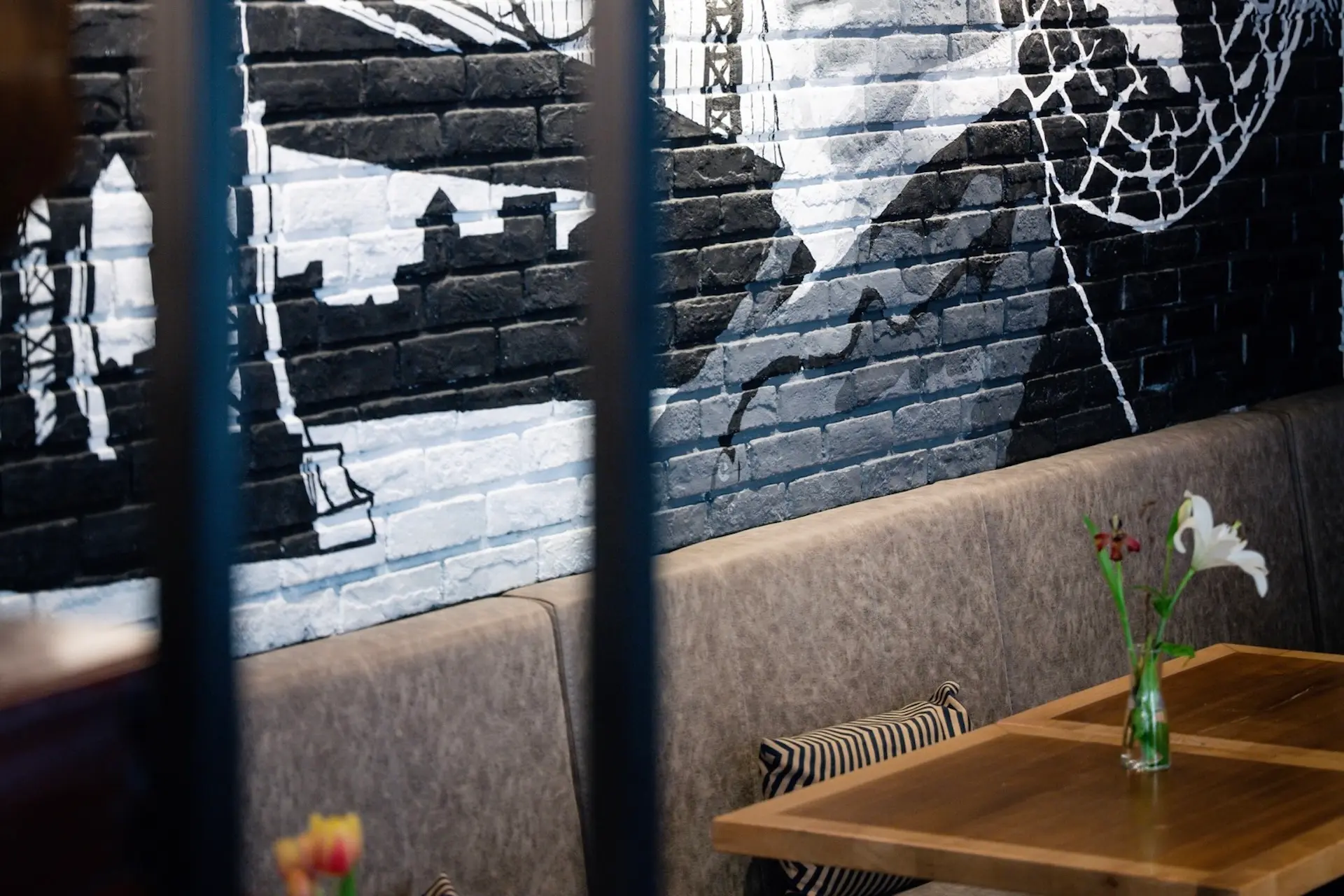 Ein Sitzbereich im Restaurant mit gepolsterten Bänken, gestreiften Kissen und einem Holztisch. Dahinter zeigt ein Wandgemälde auf einer Backsteinwand eine Stadtlandschaft in Schwarz-, Weiß- und Grautönen. Eine Vase mit Blumen steht auf dem Tisch.