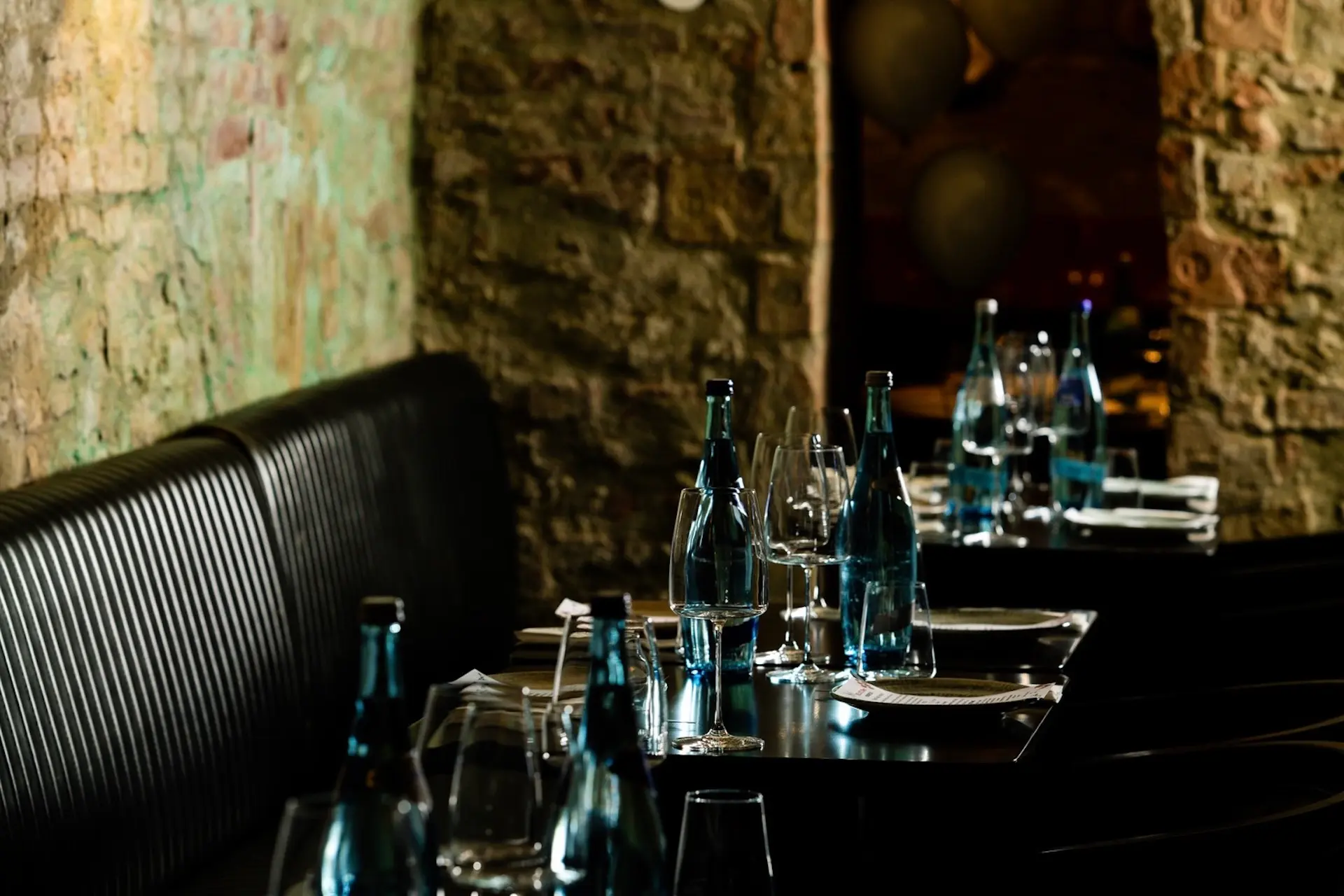 Ein schwach beleuchtetes Restaurant mit dunklen Tischen, an denen Wasserflaschen, Weingläser und Teller stehen. Der Hintergrund besteht aus einer strukturierten Steinwand und gepolsterten Sitzbänken.