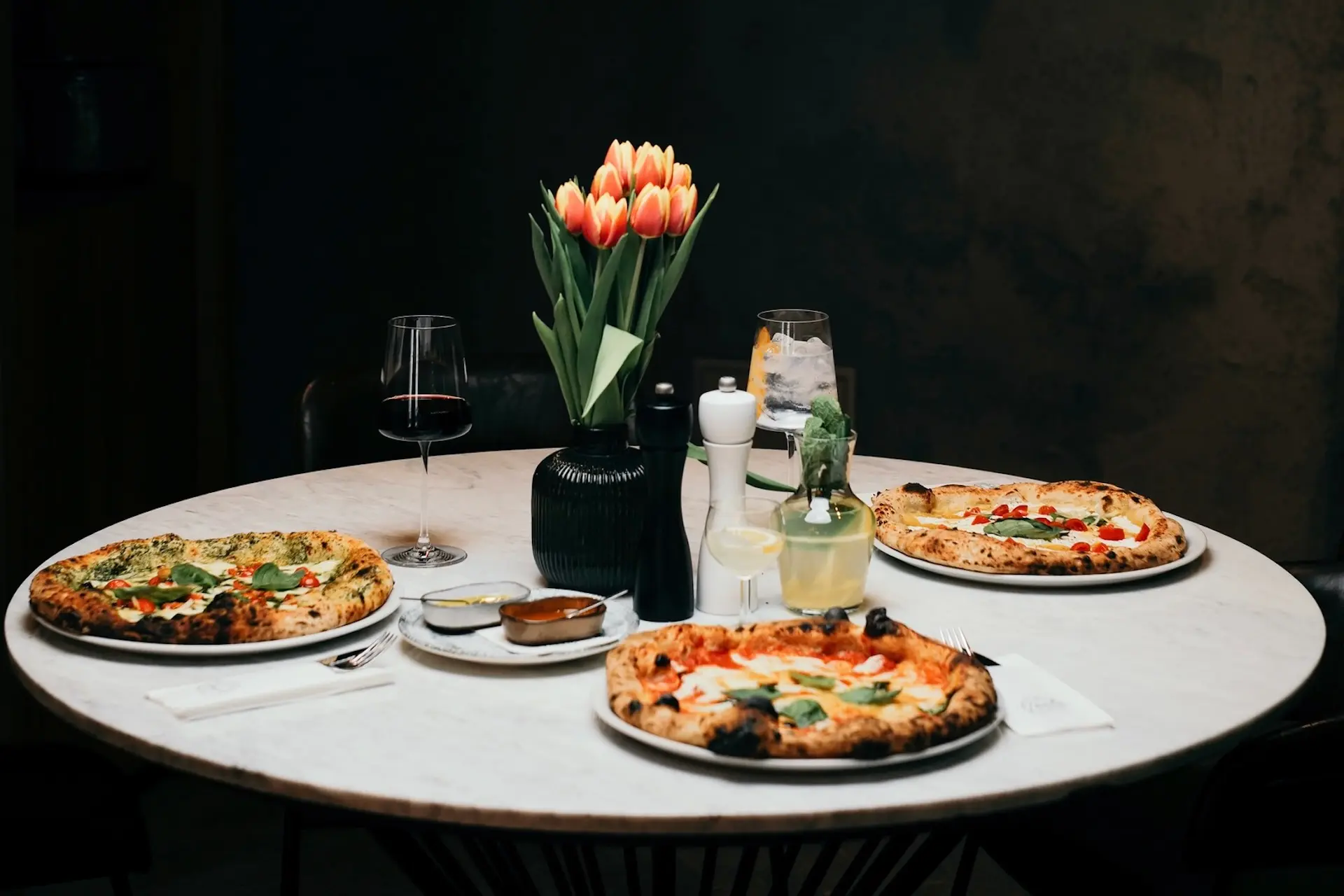 Ein runder Tisch, gedeckt mit drei Pizzen, zwei Cocktails, einem Glas Rotwein, einer Vase mit orangefarbenen Tulpen und verschiedenen Gewürzen vor einem dunklen Hintergrund.