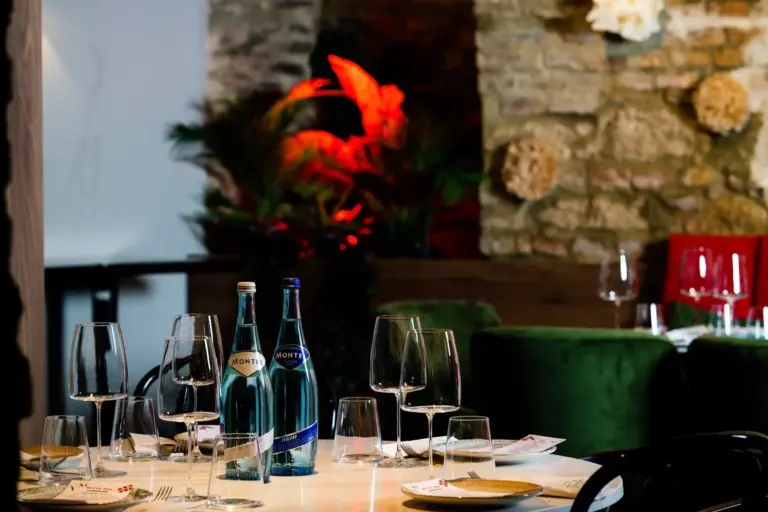 Ein Restauranttisch, gedeckt mit Weingläsern, Tellern, Speisekarten und zwei Flaschen Wasser. Im Hintergrund stehen grüne Stühle, eine Steinmauer und rot beleuchtete Pflanzen, die eine gemütliche Atmosphäre schaffen.