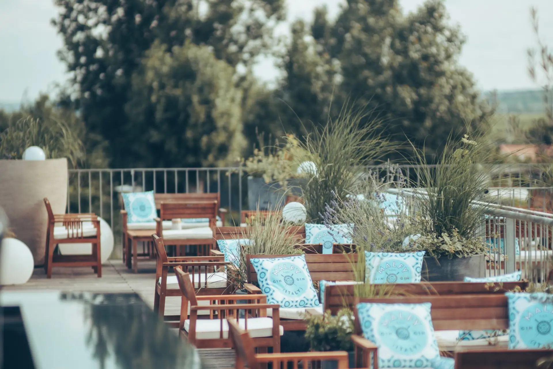 Die hölzernen Lounge-Sessel mit blau-weiß gemusterten Kissen stehen am Pool auf dem Dach, umgeben von grünen Pflanzen und Bäumen, und schaffen eine entspannte und einladende Atmosphäre.
