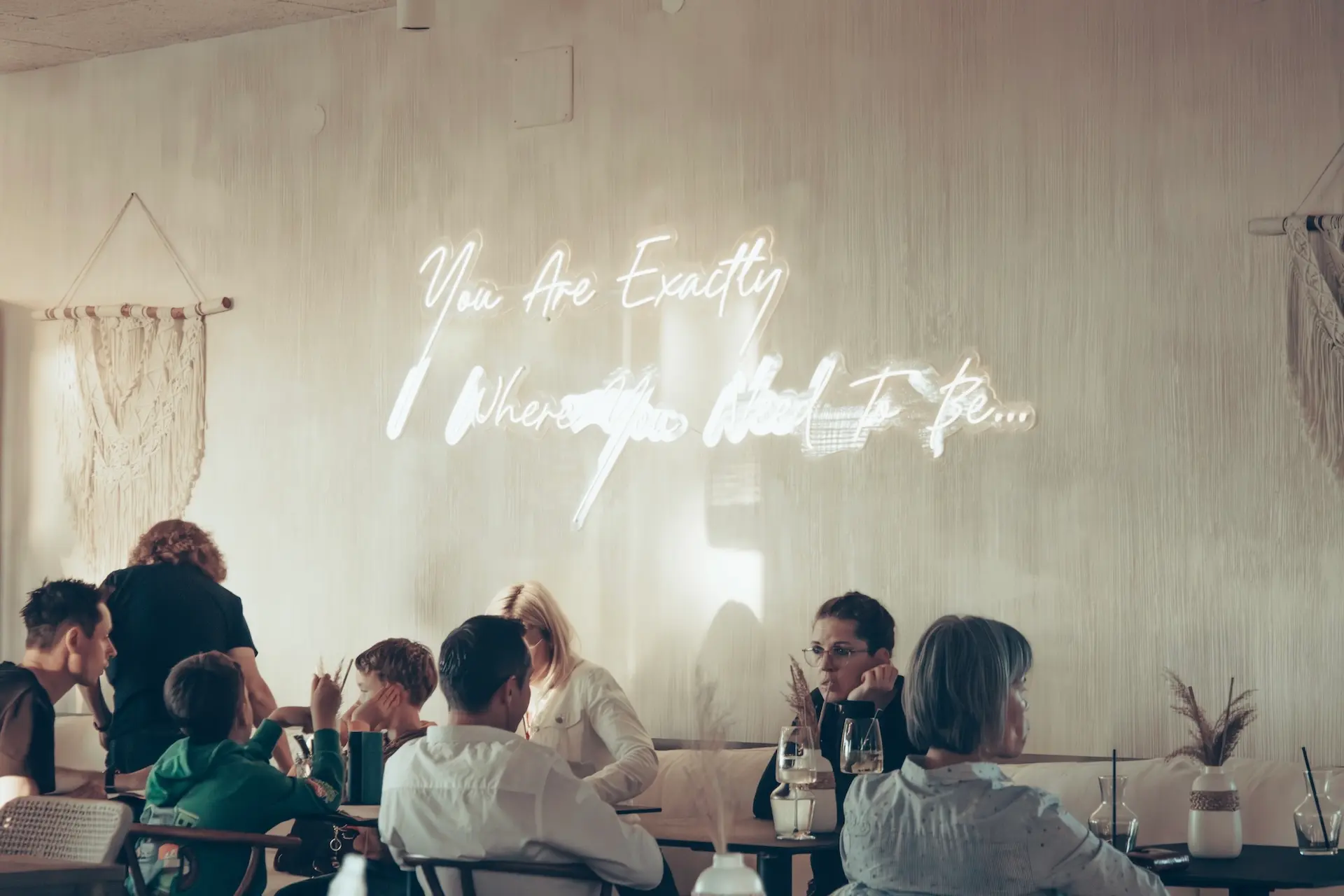Die Menschen sitzen an Tischen in einem sanft beleuchteten Café oder Restaurant, auf den Tischen stehen Getränke und Speisen. Auf einer Leuchtreklame an der Wand steht: "Du bist genau da, wo du sein sollst...
